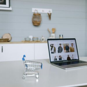 A laptop displaying an online shopping portal sits on a minimal desk with a miniature shopping cart in focus.